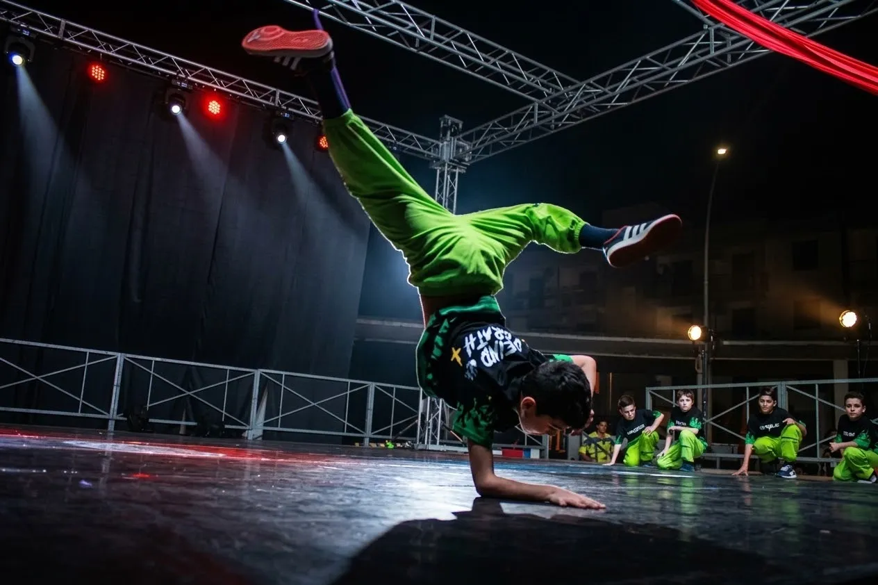 Un giovane breaker in equilibrio su una mano durante un’esibizione serale sul palco, con i compagni in tuta verde a bordo pista