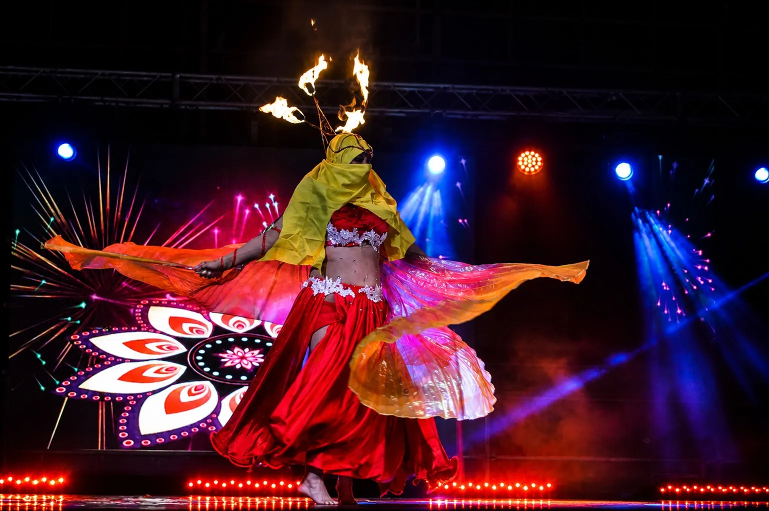 Una danzatrice orientale in scena con il volto velato e le fiamme sul capo, le ali di velo arancione aperte in una rotazione